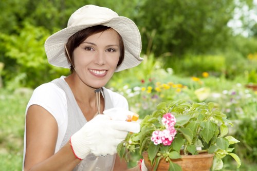 A young woman with short brown hair, wearing a wide-brimmed white hat, white gardening gloves, and a light-colored sleeveless top, is outdoors in a lush garden during daytime. She is smiling and appears to be tending to a terracotta pot filled with a flowering plant that has pink blossoms and green foliage. The background shows a well-maintained garden with a variety of plants, including shrubs and trees with dense green leaves, and colorful flowers scattered across the landscape. A neatly mowed lawn with vibrant green grass is visible in the foreground, bordered by established garden beds and a paved pathway. The scene suggests an organised outdoor space suitable for gardening and landscaping services, with natural daylight illuminating the scene, indicating a clear, bright day. This setting aligns with the local context of Battersea, where professional gardening or hedge trimming services are often requested for such well-kept gardens, reflecting a dedication to outdoor maintenance and beautification.