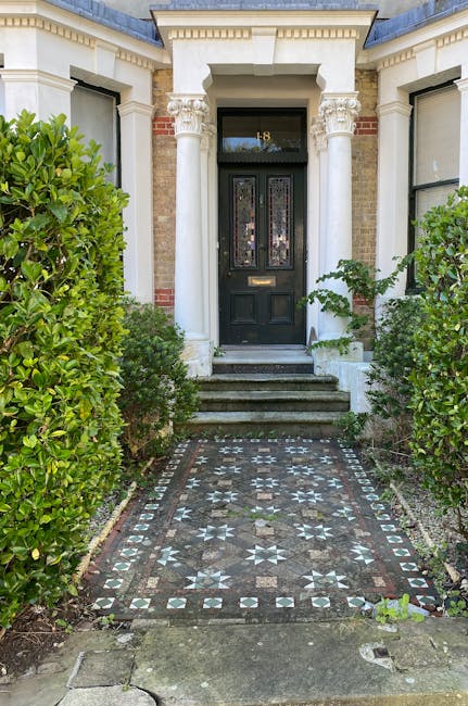 A paved front pathway leading to a black front door with decorative glass panels, flanked by white classical columns and brickwork on a residential building. The pathway features geometric patterned t