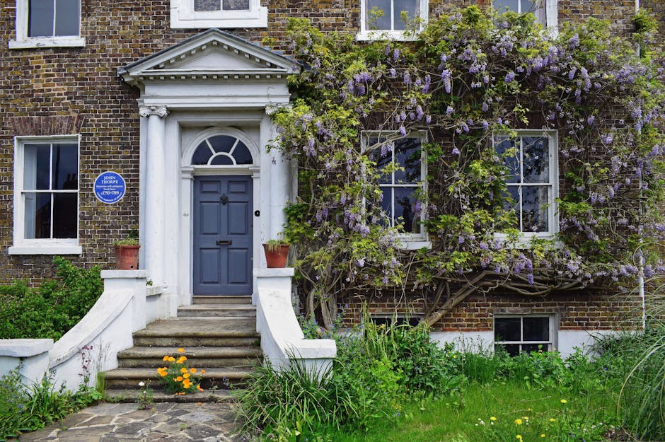 A paved front pathway leading to a black front door with decorative glass panels, flanked by white classical columns and brickwork on a residential building. The pathway features geometric patterned t