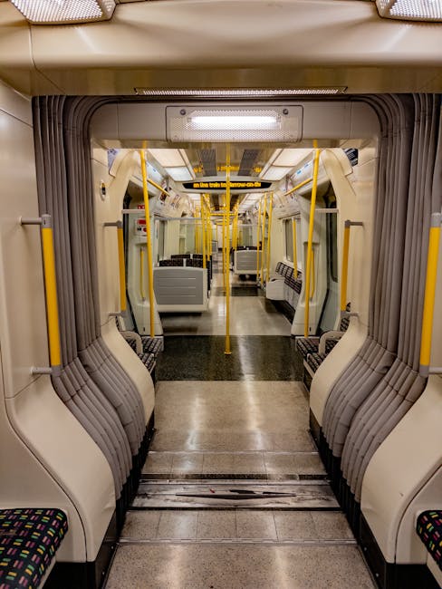 An interior view of a modern, empty train carriage featuring rows of patterned fabric seats along both sides, separated by a central aisle with a non-slip rubber mat. The beige and gray seat shells ar